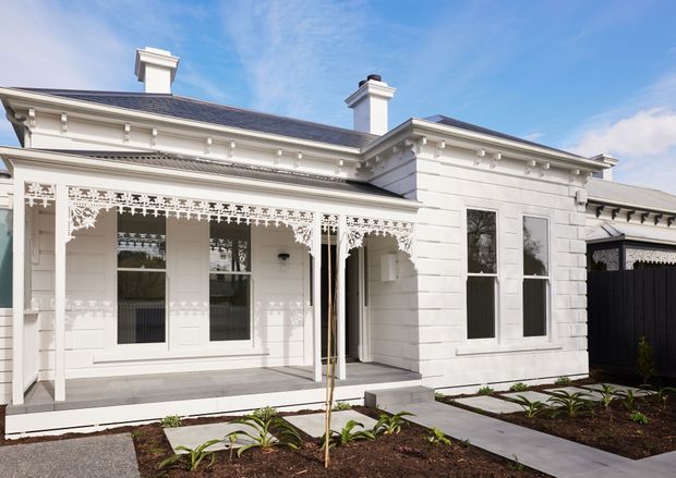 Exterior view of a restored white heritage-style residence with a detailed covered veranda, decorative lacework, classical cornices, tall sash-style windows, rendered horizontal wall bands, and multiple chimneys. The front garden contains newly planted beds and concrete paths, while a dark fence borders the property.