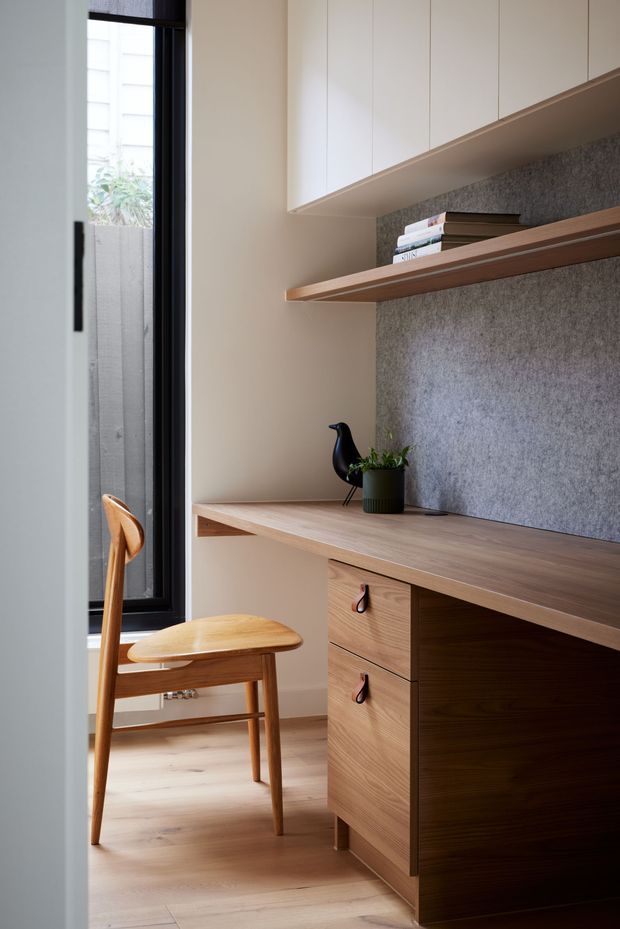 A compact, refined home office nook with a built-in timber desk, drawers, open shelving, and overhead white cabinetry. A grey textured felt-like wall panel forms the backdrop, while a warm mid-century wooden chair sits beside a tall black-framed window. The space has light timber flooring, warm neutral walls, and understated decorative styling.