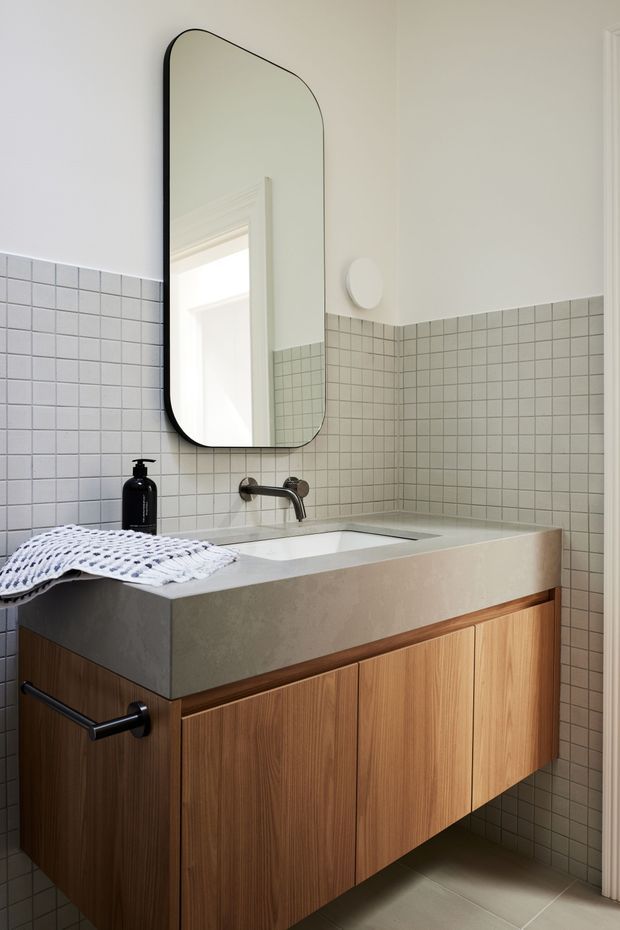 A refined contemporary bathroom vanity with a floating vertical-grain timber cabinet, thick pale concrete or engineered-stone benchtop, integrated rectangular basin, and matte black wall-mounted tapware. Small square light grey tiles cover the lower walls, while the upper walls are painted off-white. A tall rounded-corner black-framed mirror, round wall light, black soap dispenser, and folded white towel complete the compact, clean-lined space.