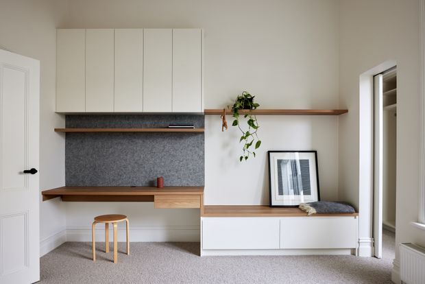 A bright, refined built-in home office and storage wall in a neutral room. The composition includes white upper cabinetry, a timber floating shelf and desk, a grey textured backsplash or pinboard surface, white lower cabinetry, and a small timber stool. A framed print and trailing plant add understated decoration, while an open doorway reveals additional storage.