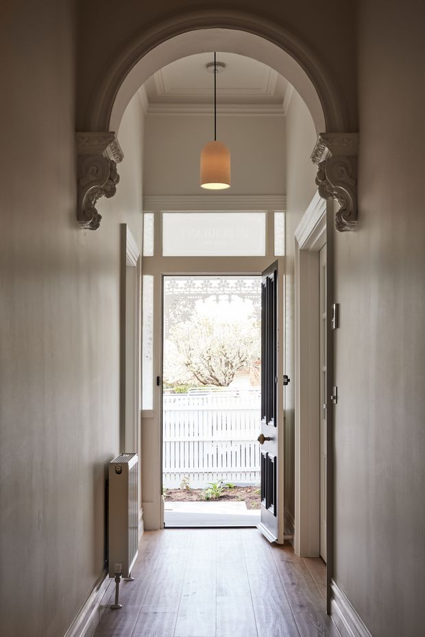A narrow, elegant residential entry hallway viewed toward an open front door. The corridor has warm grey painted walls, a timber floor, white decorative skirting and ceiling mouldings, and a large ornate arched opening with carved classical corbels. A pendant light hangs centrally, with a radiator along the left wall and built-in shelving or cabinetry on the right. Daylight enters through the open glazed door and fanlight, creating a refined heritage-style entrance.
