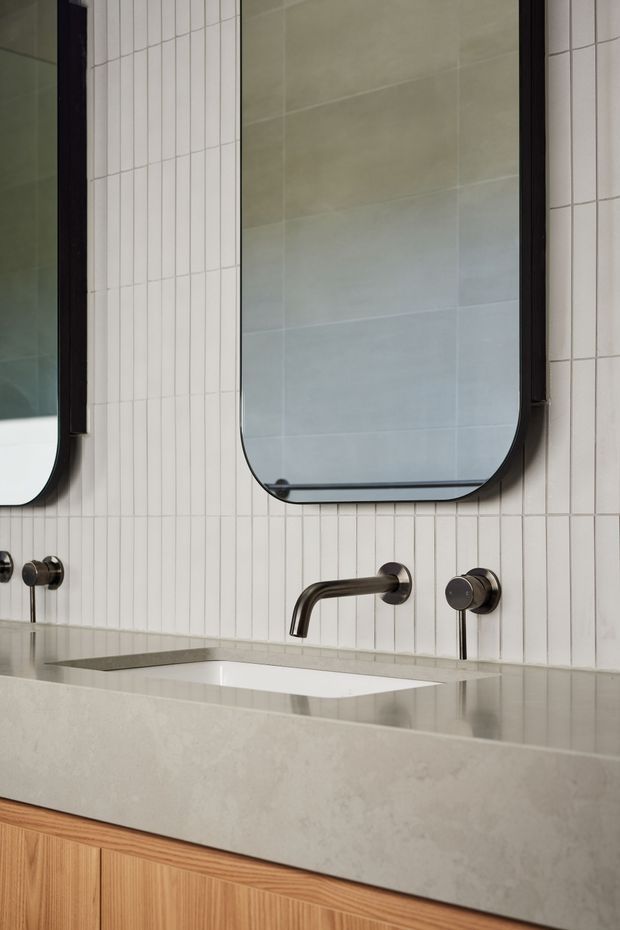 Close-up view of a contemporary bathroom vanity with a pale stone benchtop, integrated white basin, wall-mounted dark metal tapware, and vertically arranged white wall tiles. Rounded-rectangle mirrors with dark frames hang above the vanity, while warm timber cabinetry provides contrast below.