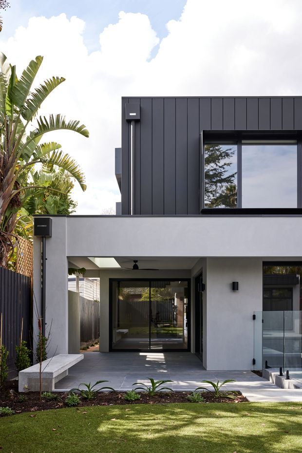 Exterior view of a contemporary two-storey home with a dark vertically panelled upper facade, smooth light-rendered lower level, large black-framed glazing, and a covered entry or outdoor living area. A landscaped front garden, low concrete bench, glass balustrade, boundary fencing, and tropical planting are visible.