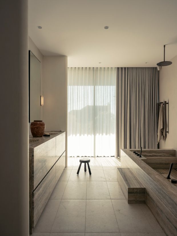 Minimal, warm-toned contemporary bathroom with a freestanding double vanity and broad stone counter on the left, a large built-in stone-clad bathtub or bathing platform on the right, a wall-mounted shower, floor-to-ceiling sheer and blackout curtains, and large pale stone floor tiles. Natural light filters through the window, creating a calm, refined atmosphere.
