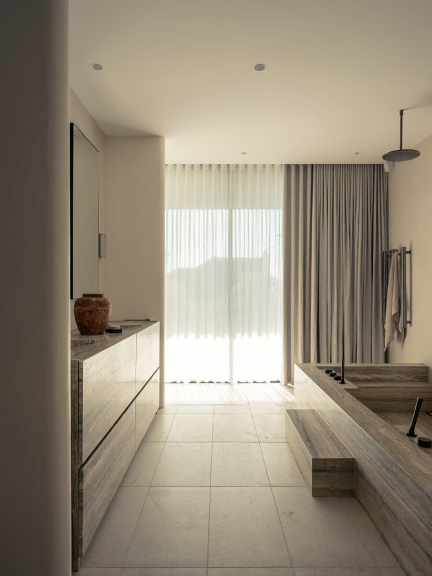 A refined contemporary bathroom viewed from the entry, featuring a freestanding-style built-in stone bathtub and integrated timber-look vanity cabinetry. Large pale floor tiles, warm neutral walls, layered sheer and grey drapery, recessed ceiling lights, and a walk-in shower with a dark ceiling-mounted showerhead create a calm, softly lit interior. A bright glazed opening is visible at the far end.