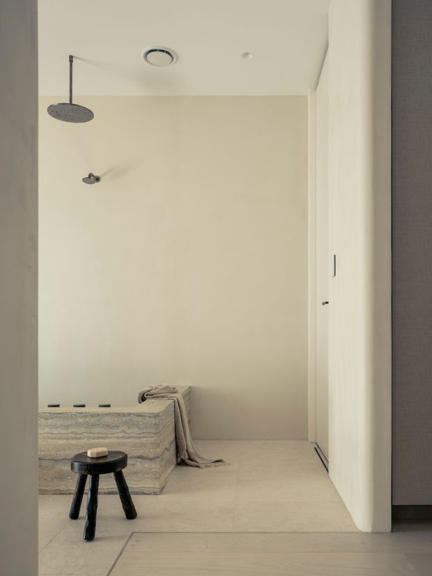 Minimal, warm-neutral bathroom with a walk-in shower, large round rainfall showerhead, pale stone or tiled walls and floor, and a sculptural freestanding bathtub partially visible at the left. A small dark stool sits in the foreground, while a concealed doorway or storage panel appears on the right. The space is softly lit and uncluttered.