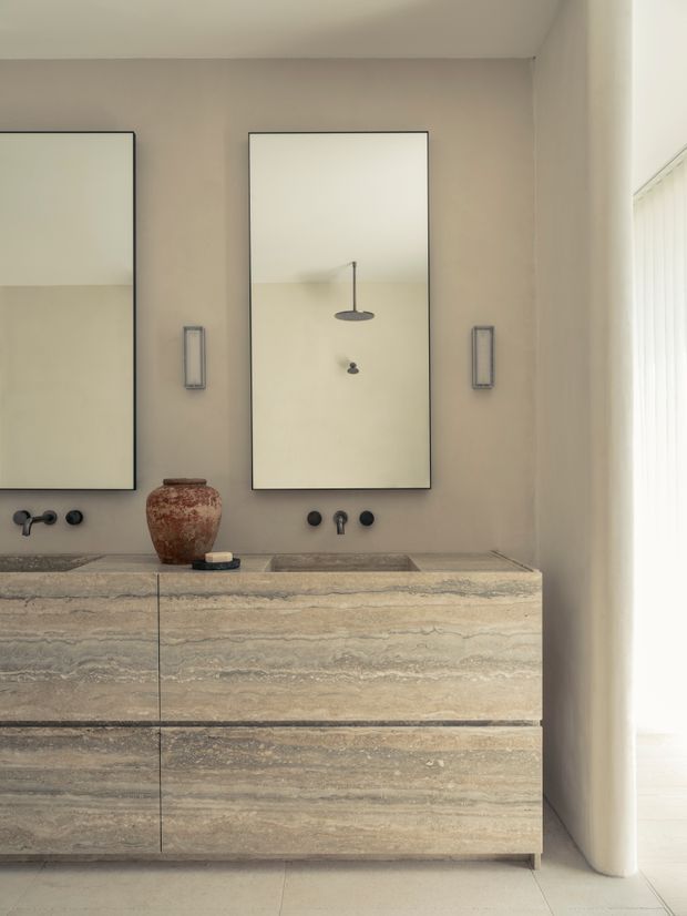 A refined bathroom vanity area with a double travertine stone vanity, integrated basins, dark wall-mounted tapware, two large rectangular mirrors, and slim wall sconces. Neutral taupe walls, warm stone surfaces, and sheer vertical blinds create a calm, softly lit interior.
