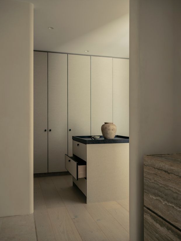 A softly lit minimalist interior showing a compact built-in cabinetry zone, viewed through a doorway or opening. Full-height pale cabinetry lines the rear wall, while a central island or console has a dark stone benchtop, integrated drawers, and a ceramic vessel. Pale timber flooring, warm off-white walls, and a partial stone-clad surface at right create a restrained, calm material palette.