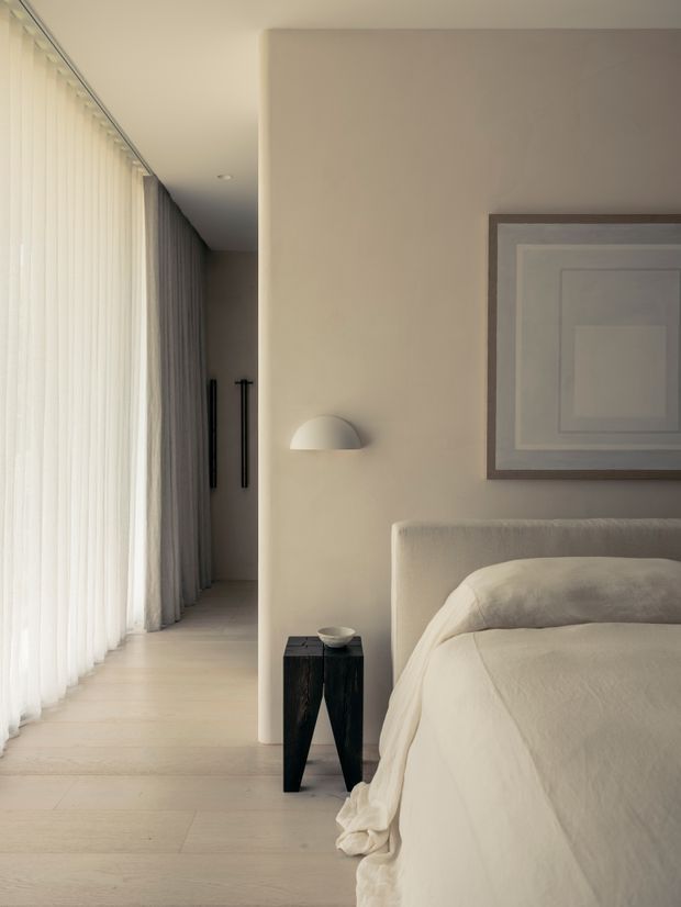 A softly lit, neutral-toned bedroom with a partially visible upholstered bed, layered cream bedding, a dark sculptural bedside table, wall-mounted dome light, and framed artwork. Floor-to-ceiling sheer curtains and heavier drapes line the adjacent passage, creating an airy, calm interior with pale timber flooring and minimalist detailing.