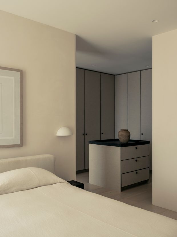 A softly lit bedroom with a low upholstered bed in the foreground, warm beige walls, pale bedding, and a minimalist built-in wardrobe and dressing island at the rear. The island has a dark stone-like benchtop and integrated drawers, with a ceramic vessel placed on top. A simple wall-mounted half-dome light and framed artwork complete the calm, neutral interior.