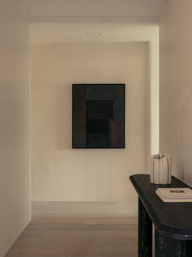 A quiet, minimalist interior passage or entry space with warm off-white plaster walls, pale timber flooring, and recessed ceiling lights. A dark abstract artwork hangs on the rear wall, while a dark console table with a sculptural light-coloured vase and books occupies the right foreground.