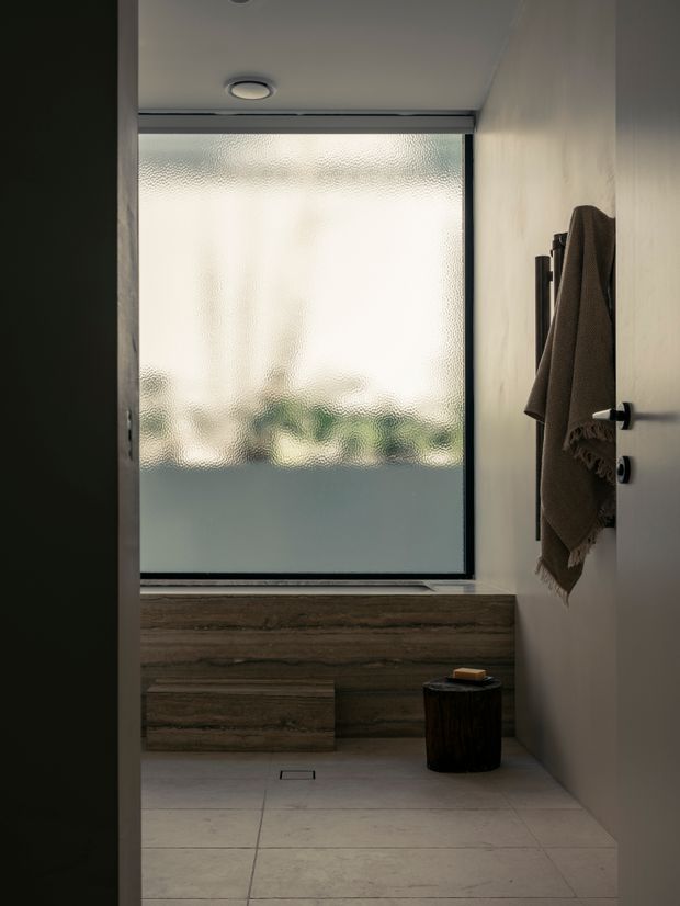 A compact, minimalist bathroom viewed through an open doorway. The room features a freestanding or built-in stone-clad bathtub beneath a large frosted window, pale large-format floor tiles, smooth light walls, a dark towel rail with a draped brown towel, and a small dark timber stool. Soft diffused daylight creates a calm, understated atmosphere.