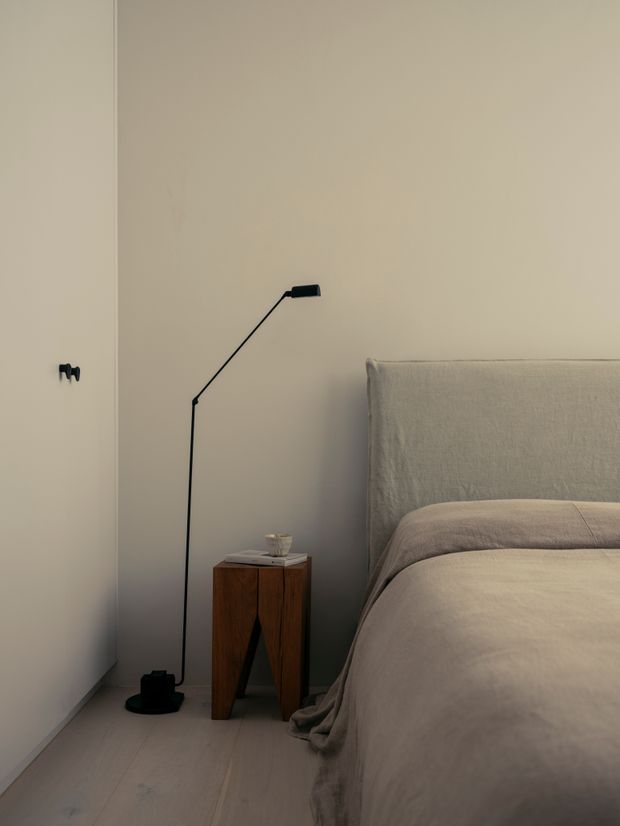 A softly lit minimalist bedroom corner featuring a neutral upholstered bed with layered taupe bedding, a slim black floor lamp, and a small dark timber bedside table. Pale walls and light timber flooring create a calm, understated atmosphere.