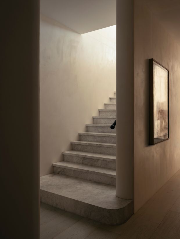 A softly lit interior stairway viewed through a narrow opening. Broad pale stone or terrazzo-clad steps rise along a plastered wall toward a bright overhead opening, with a rounded stair base and cylindrical column in the foreground. Warm timber flooring and a framed artwork on the adjacent wall add understated detail.