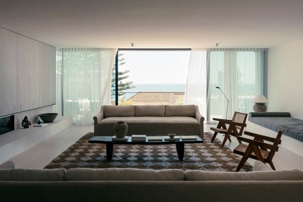 A bright, refined mid-century-inspired living room with a large sectional sofa facing a low black coffee table and additional lounge seating. Floor-to-ceiling glazing with sheer curtains frames a clear ocean view, while pale built-in cabinetry and a patterned area rug add texture to the otherwise neutral interior.