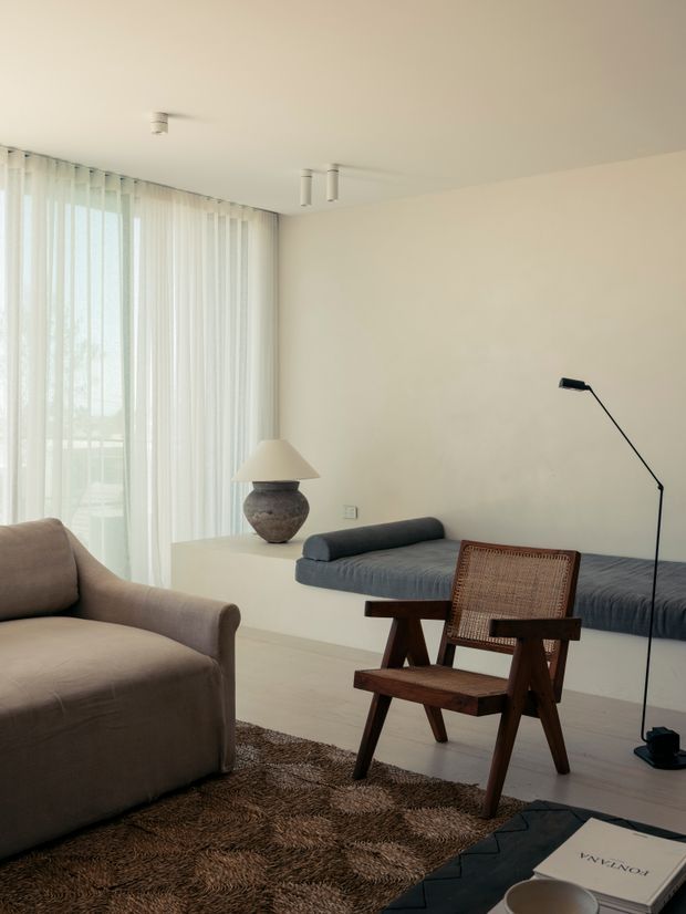 A softly lit minimalist living room with floor-to-ceiling sheer curtains, a low built-in daybed or sofa along the wall, a sculptural table lamp, a plush upholstered armchair, a woven wooden lounge chair, and a dark shag rug. The neutral palette and simple furnishings create a calm, refined atmosphere.