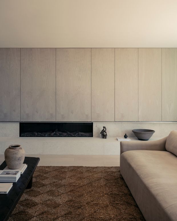 Minimalist contemporary living room with a long built-in timber-panelled wall, recessed linear fireplace, pale textured hearth, neutral upholstered sectional sofa, dark low coffee table, ceramic vessel and a brown textured rug. The restrained palette and concealed joinery create a calm, refined atmosphere.