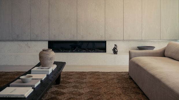 Minimalist living room with a long, low black coffee table, neutral upholstered sectional sofa, textured brown rug, ceramic vessel, and a recessed linear fireplace set into a pale stone or plaster wall beneath full-height light timber wall panelling.