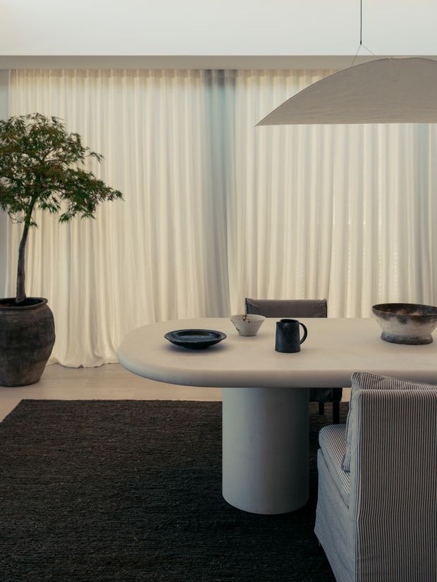 A refined dining area with a sculptural oval table on a single cylindrical pedestal, upholstered dining chairs, a large neutral pendant light, sheer floor-to-ceiling curtains, dark textured rug, ceramic tableware, and a potted indoor tree. The room has a restrained, contemporary minimalist aesthetic with soft neutral tones and organic forms.