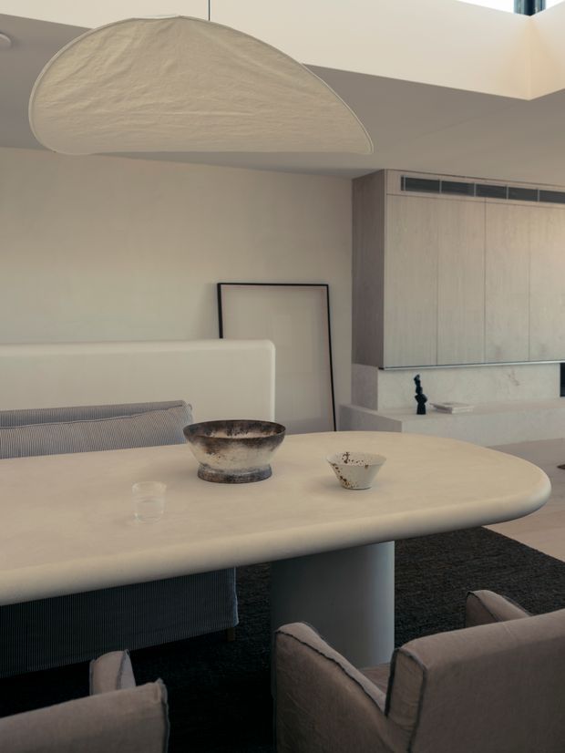 A refined contemporary dining area with a large rounded-edge cream dining table, upholstered dining chairs, a sculptural pendant light, and a minimalist built-in cabinet with integrated ventilation. Neutral plaster walls, a dark rug, subtle artwork, and ceramic vessels create a calm, softly textured interior.