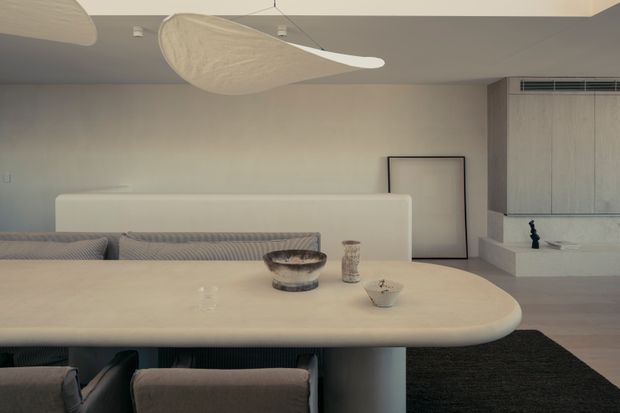 A refined open-plan living and dining area with a rounded light stone or solid-surface dining table, upholstered dining chairs, and a low-profile sofa behind it. The room features pale neutral walls, light timber built-in cabinetry, a simple linear fireplace or plinth, dark flooring accents, and sculptural organic pendant lights. The overall composition is calm, minimalist, and softly textured.