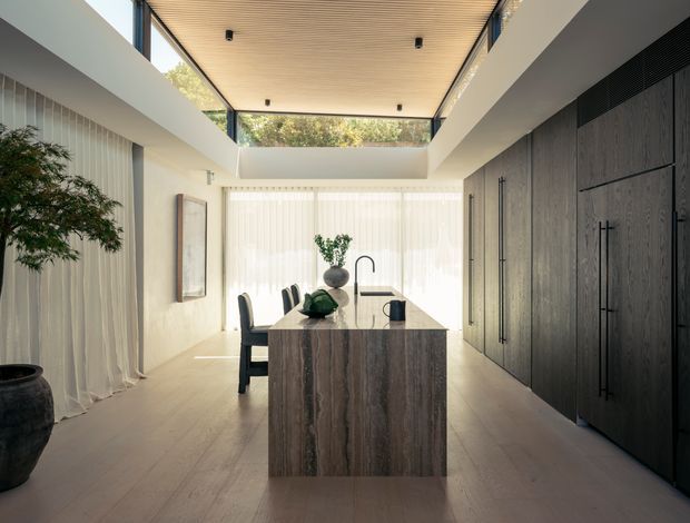 A refined contemporary kitchen viewed from the living area, featuring a large dark stone island with an integrated sink and black tap, tall dark timber cabinetry, pale timber flooring, sheer curtains, and a dramatic double-height ceiling with a warm timber-lined skylight or clerestory opening. Natural light fills the space and highlights the restrained, minimalist material palette.