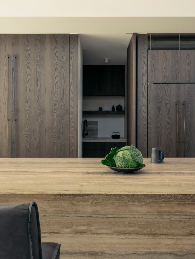 A refined contemporary kitchen viewed across a substantial timber island bench. The kitchen features full-height dark smoked timber cabinetry, integrated vertical handles, a concealed pantry or preparation area, dark shelving, and a black sink tap. A bowl of cabbages and a dark metal jug sit on the island, with a partial upholstered bar stool in the foreground. The palette is warm and subdued, dominated by natural timber and charcoal tones.