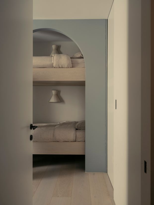 A softly lit compact bunk bedroom viewed through a doorway, featuring two built-in bunk beds with rounded arch openings, pale timber platforms and flooring, warm neutral bedding, and small wall-mounted fabric-shaded lights. A tall pale built-in cupboard occupies the right side of the frame, creating a calm, minimalist children’s sleeping space.