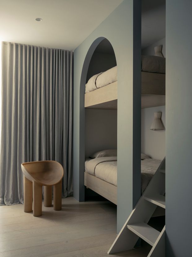 A compact, softly lit bunk bedroom with built-in bunk beds framed by deep blue-grey joinery and rounded arch openings. Pale bedding, full-height grey curtains, light timber flooring, a sculptural wooden stool, and an integrated ladder create a calm, contemporary children's sleeping space.