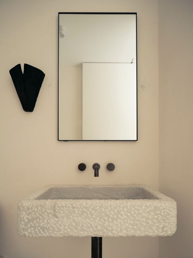 A compact, minimalist bathroom vanity with a textured white rectangular basin, wall-mounted black tapware, and a large black-framed mirror. The warm beige wall and sculptural black wall accent create a restrained, refined look.