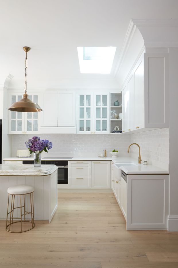Bright, refined white kitchen with full-height shaker-style cabinetry, glazed display cupboards, a marble-look island benchtop, white subway-tile splashback, pale timber flooring, brass pendant lighting and a brass gooseneck tap. A skylight brings additional natural light into the space, while traditional mouldings and panelled joinery add character.