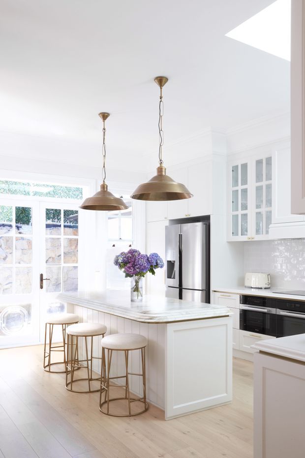 Bright, refined kitchen with a large central island, white shaker-style cabinetry, glass-front wall cabinets, stainless-steel appliances, glossy white subway-tile splashback, pale timber flooring, and a light stone island benchtop. Three stools sit at the island beneath oversized brass pendant lights. Glazed doors and windows bring in abundant natural light, with a bouquet providing a small colour accent.