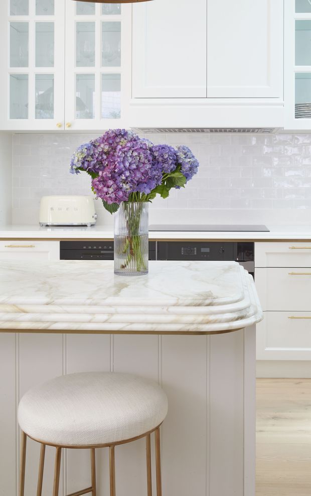 Bright, refined kitchen interior featuring white shaker-style cabinetry with glass-front doors, glossy white subway-tile splashback, integrated black appliances, pale timber flooring, and a large island with a rounded, veined white marble benchtop and paneled base. A light upholstered stool and a vase of purple hydrangeas add softness and colour.