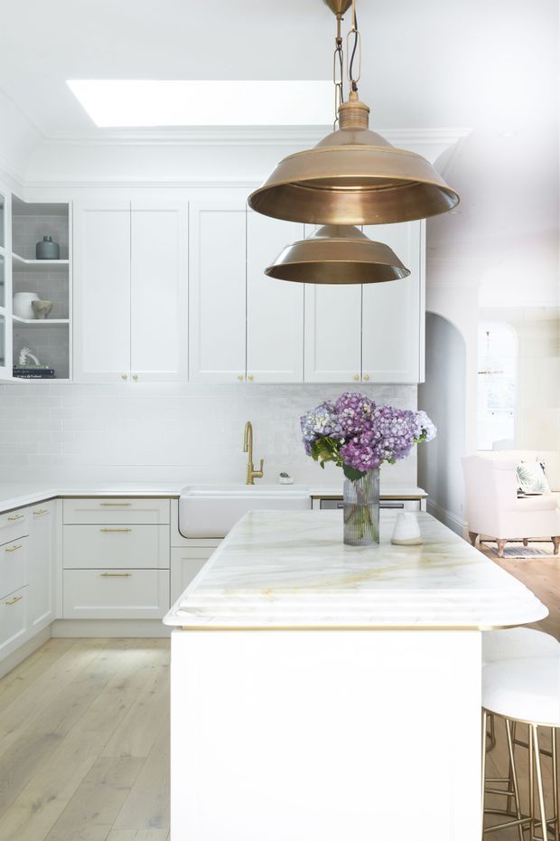 Bright, white traditional-style kitchen with a large central island, pale stone benchtop, white shaker cabinetry, brass hardware, farmhouse sink, white tiled splashback, and paired brass pendant lights. A skylight illuminates the room, while an arched opening reveals part of an adjacent living area. Light timber flooring and a vase of flowers add warmth.