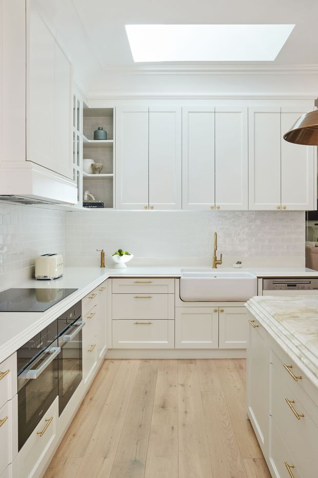 Bright, refined white kitchen with traditional Shaker-style cabinetry, brass hardware, a farmhouse apron-front sink, pale timber flooring, and a light textured brick-pattern backsplash. A large skylight provides abundant natural light, while white stone benchtops and integrated appliances create a clean, classic fit-out.