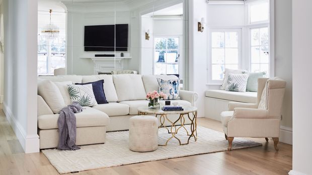 Bright, refined living room with a large cream sectional sofa, upholstered armchairs, a round glass coffee table with a decorative gold-toned base, and a soft area rug over light timber flooring. White wall panelling, tall multi-pane windows, detailed ceiling mouldings, and a built-in media wall create a classic coastal-inspired interior.
