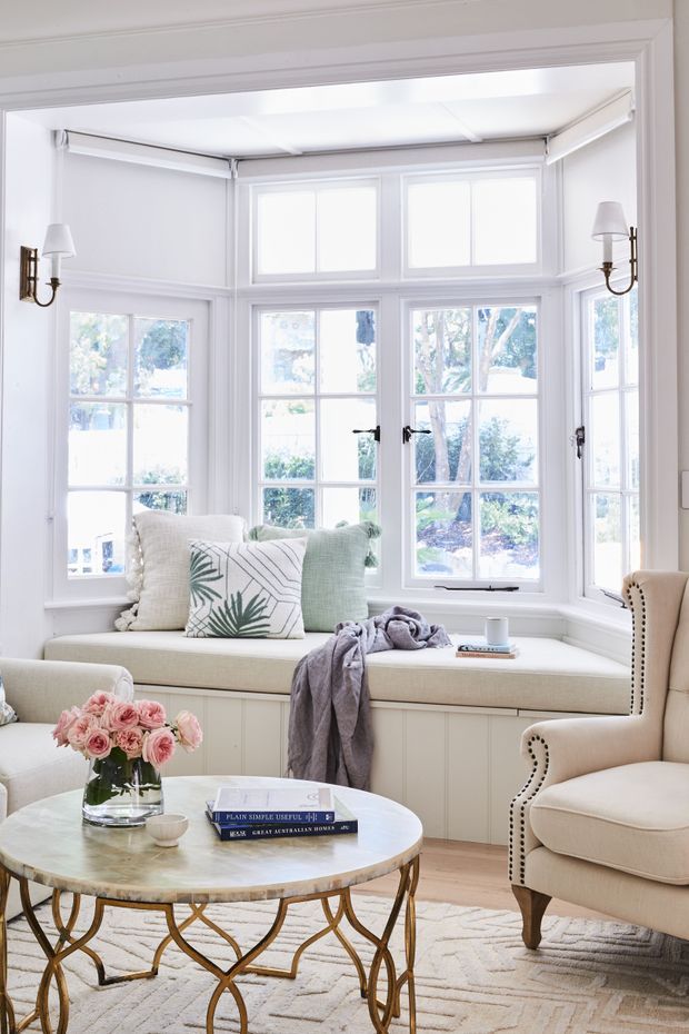 Bright, elegant sitting room centred on a white-painted bay window with built-in upholstered window seating and abundant multi-pane glazing. The room includes a cream armchair with nailhead trim, a round marble-topped coffee table with a brass-toned sculptural base, pale upholstered cushions, flowers, and a light patterned rug. The overall look is refined, traditional, and airy.