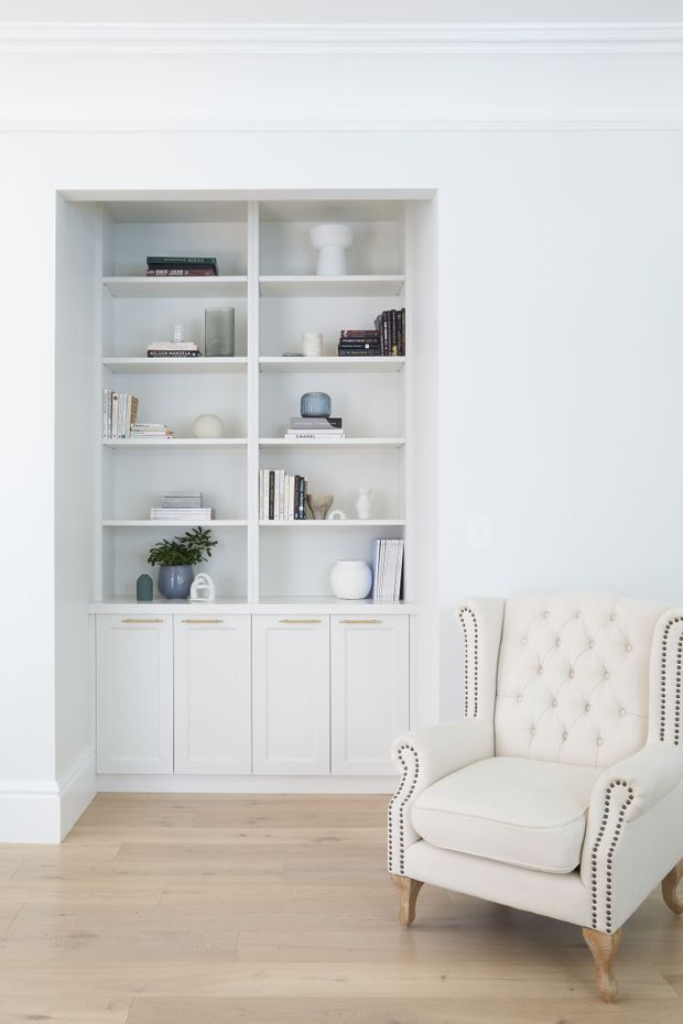 A bright, refined interior featuring a built-in white bookcase with open shelving and lower paneled cabinetry, styled with books and minimal decorative objects. A plush cream tufted wingback armchair with nailhead trim sits in the foreground on pale timber flooring. White walls and classic ceiling and wall mouldings create a clean, elegant reading or living area.