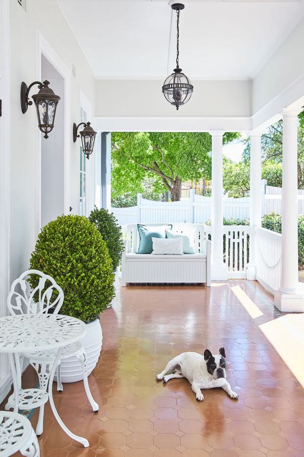 Bright covered residential porch/veranda with white columns, balustrades, fencing, and trim. The space features terracotta hexagonal tile flooring, ornate dark metal wall lanterns and pendant lighting, white wrought-iron café furniture, clipped topiary plants, and a built-in white outdoor daybed with pale blue cushions. A small dog is present on the floor.