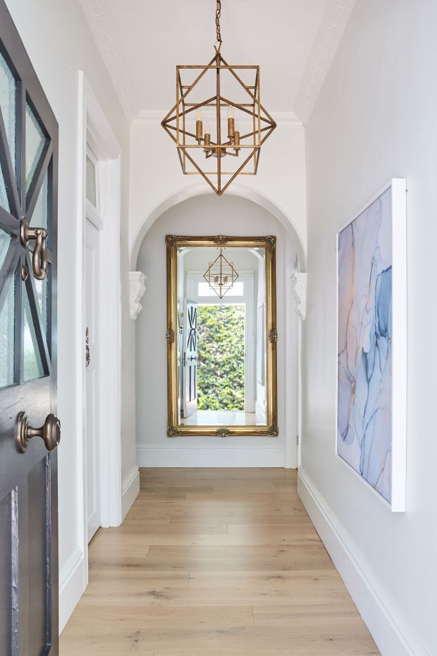 A bright, narrow residential hallway or entry corridor with white walls, decorative ceiling mouldings, arched detailing, pale timber flooring, and a large ornate gold-framed mirror at the far end. A geometric brass pendant light hangs centrally, while a dark glazed entry door and an abstract blue-toned artwork add contrast. The mirror reflects the pendant and an open doorway leading toward greenery.