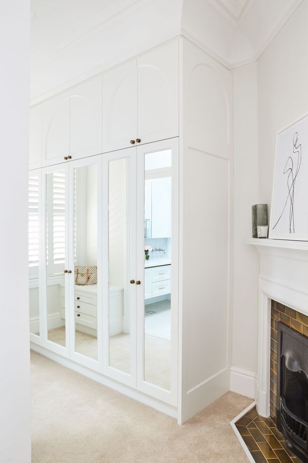 Bright, refined bedroom interior featuring a large built-in white wardrobe with mirrored doors and arched decorative panel detailing. A white fireplace with a dark tiled hearth is visible at right, while mirrored wardrobe panels reflect a pale bathroom or dressing area. The room has high decorative cornices, pale carpet, white walls, and brass-toned hardware.