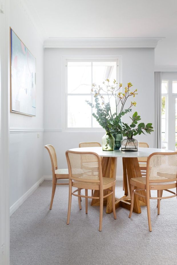 Bright, airy dining room with white walls, decorative wall moulding and trim, large white-framed windows, and a round light timber dining table surrounded by woven cane chairs. A large floral artwork and a green glass vase with leafy branches add soft colour, while pale carpet creates a calm, contemporary setting.