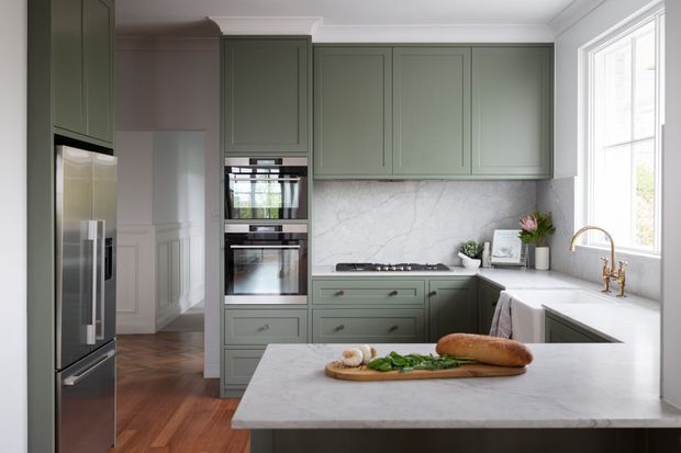 A refined kitchen with muted sage-green shaker cabinetry, integrated double ovens, a large island with a pale stone benchtop, and a matching stone splashback. The space features dark timber flooring, white walls and ceiling moulding, a stainless-steel refrigerator, brass tapware, and abundant natural light from a large window.
