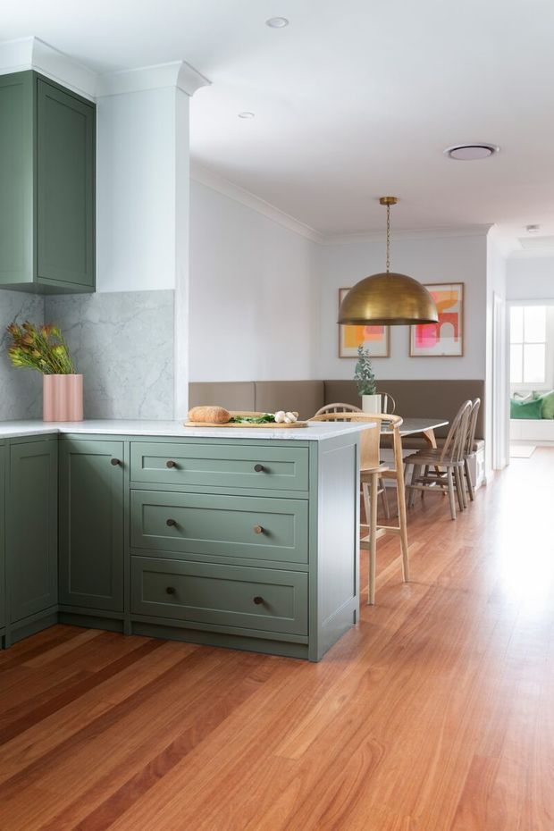 Bright open-plan kitchen and dining area with a muted sage-green island and cabinetry, pale marble-look splashback, warm timber flooring, and a compact dining nook. A large brass dome pendant hangs above the dining table, with framed abstract artwork and upholstered bench seating along the wall. White walls, decorative cornices, and recessed ceiling lights create a refined, welcoming interior.