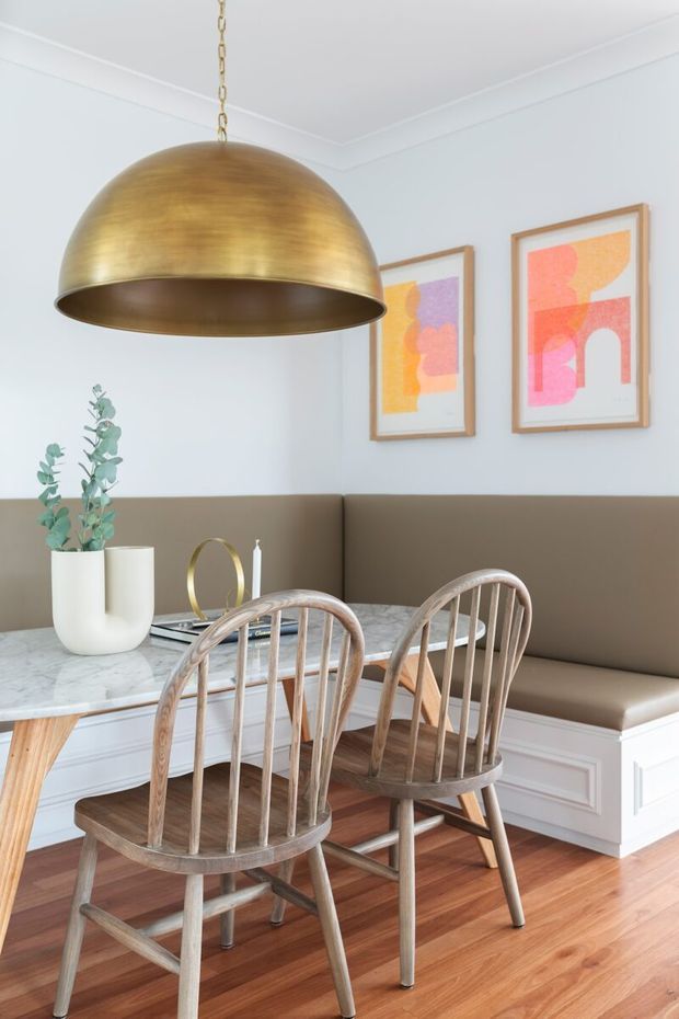 Bright, refined dining nook with a marble-topped table, curved wooden spindle-back chairs, and a built-in taupe upholstered banquette. White panelled lower walls and light grey upper walls are complemented by colourful abstract framed artwork, a large brushed brass dome pendant, and warm timber flooring.