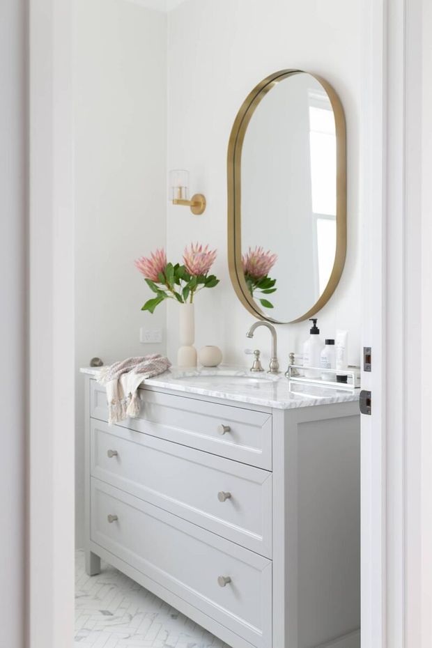 Bright, compact bathroom vanity featuring a pale grey-white paneled cabinet with drawers, a white marble-look benchtop, polished chrome tapware, and a large rounded-arch mirror with a brass frame. White patterned floor tiles, a wall-mounted light, and simple floral styling create a refined, fresh interior.