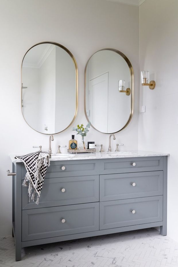 Bright, refined bathroom featuring a large grey double vanity with paneled cabinetry, a white marble-look benchtop, twin undermount basins, polished metal tapware, and two tall arched brass-framed mirrors. White herringbone floor tiles, pale walls, brass wall sconces, and restrained accessories create a clean classic look.