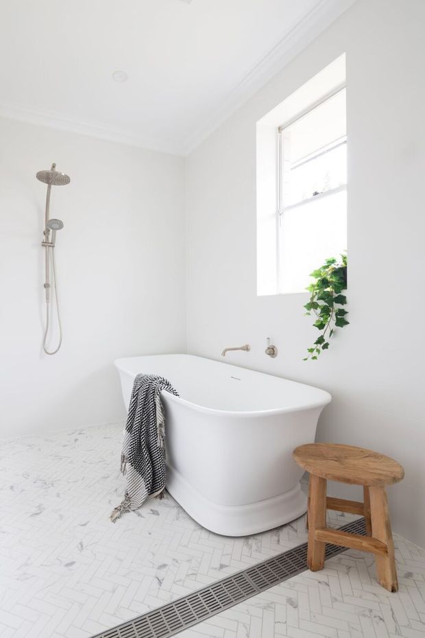 Bright, airy white bathroom featuring a freestanding bathtub, wall-mounted shower, chrome fixtures, a large window, white herringbone floor tiling, linear floor drain, and a small timber stool with trailing greenery.