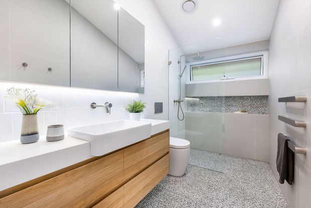 Bright contemporary bathroom with a floating timber vanity, white vessel basin, wall-to-wall mirror, wall-mounted tapware, toilet, and a walk-in shower. The shower features pale wall tiles, a horizontal mosaic feature band, a frameless glass screen, and a high awning window. The floor is finished in small-speckled terrazzo-style tiles, with minimalist chrome and dark grey accessories.