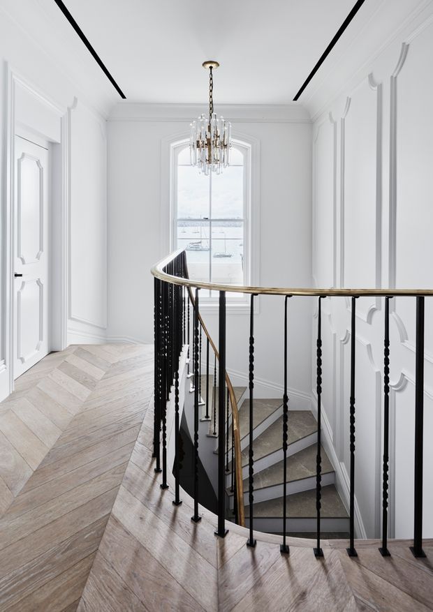 Elegant interior landing and stairwell with ornate white wall panelling, a curved black metal balustrade with brass-toned handrail, pale herringbone timber flooring, and a crystal chandelier. A tall arched window provides natural light and views beyond.